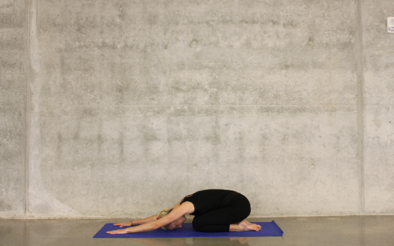 A woman performs a stretch on a blue yoga mat on the floor, in front of a concrete wall. 