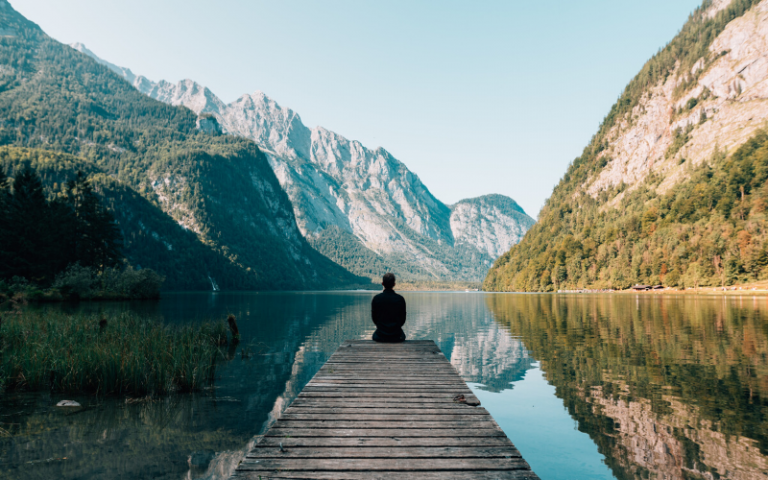 Person sitting on a wooden bridge looking out to water and mountains