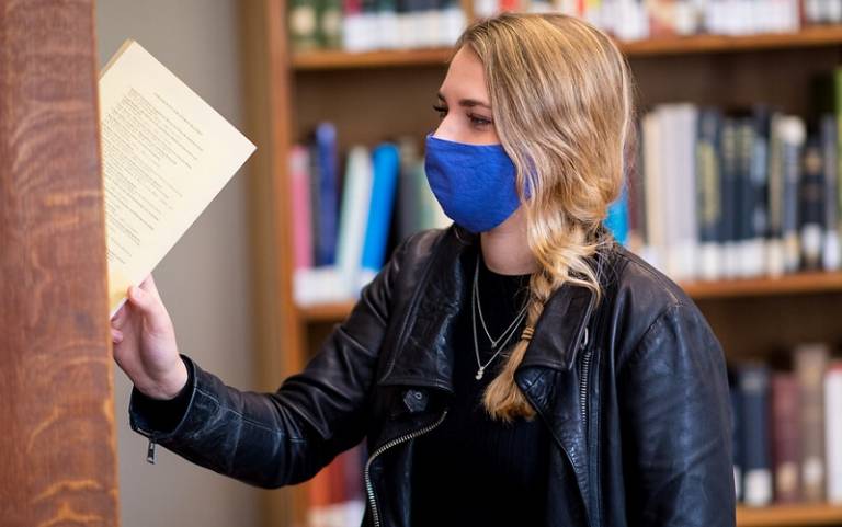 A student wearing a facemask places a text back into the shelves of a library