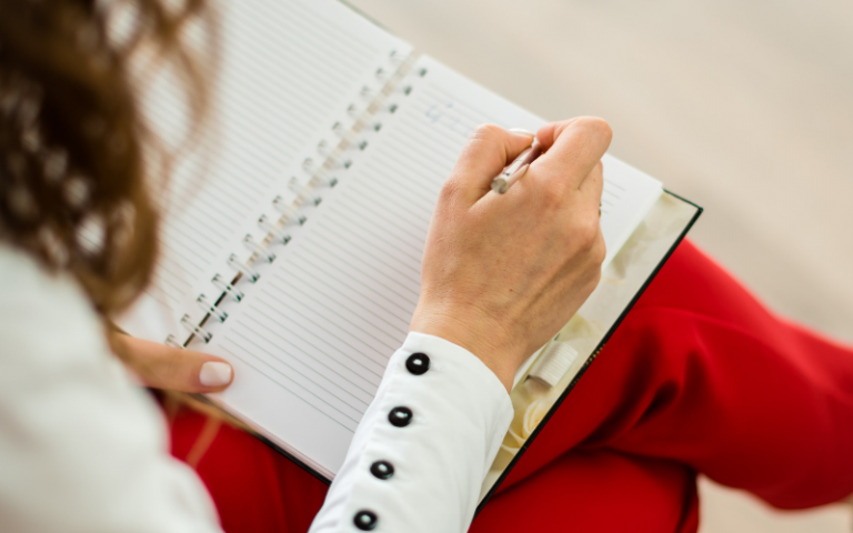 Person in white blouse with black buttons on the cuff and red trousers, sitting and writing in their planner.