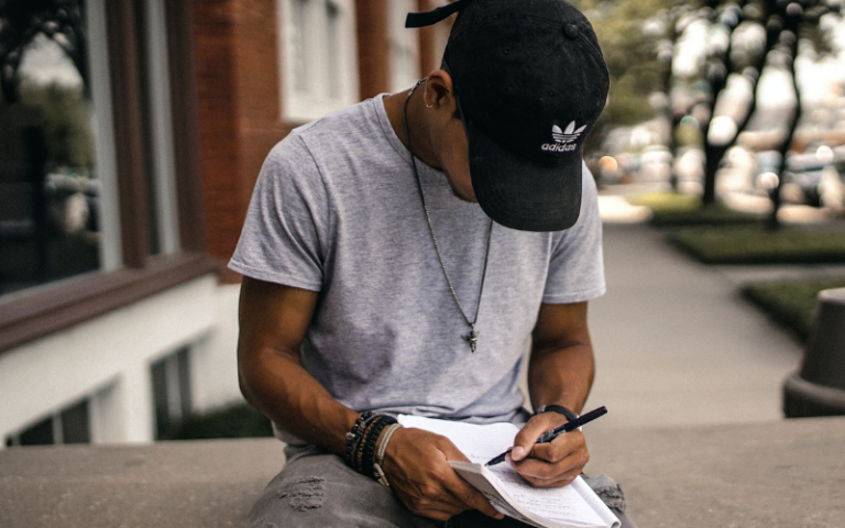 Student wearing a baseball cap sitting on a wall writing in a notebook