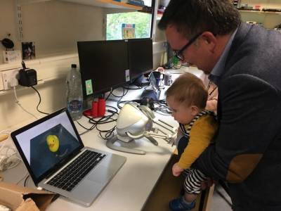 Academic holds up child to view a rubber duck on a laptop screen