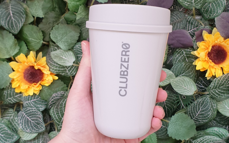 Hand holding a reusable coffee cup with the words 'Club Zero' in front of a green background with flowers