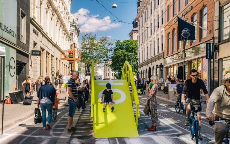 An image of a city street closed to cars, with people cycling and a child playing on a slide.