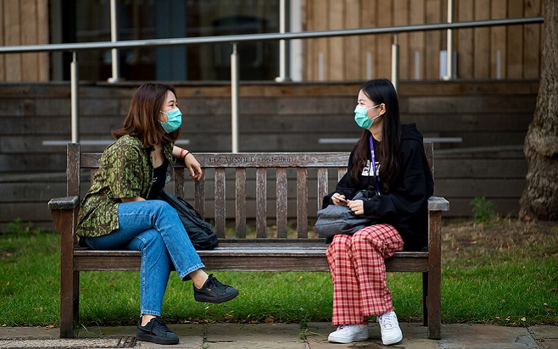 Two students wearing masks chatting on a park bench