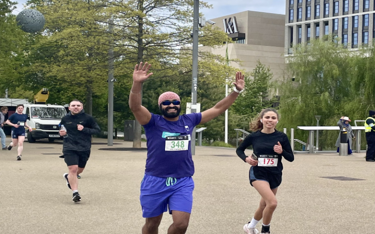 A runner raises his hands in a cheer as he runs across Queen Elizabeth Olympic Park with the V&A East in the background