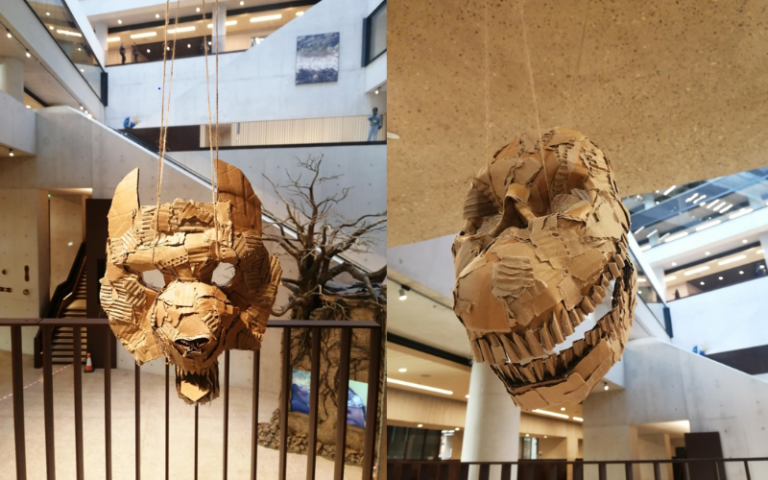 Two striking masks made from cardboard materials with animal like features hang from a terrace overlooking the atrium of UCL East's Marshgate building