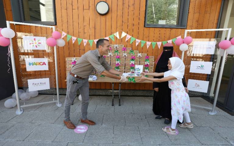 Head teacher reaches holds out a box containing a laptop to a young primary aged pupil and her mother with a background of celebratory balloons