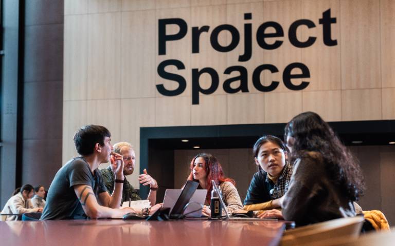A group of people sit at a table with laptops beneath a sign that reads 'Project Space'