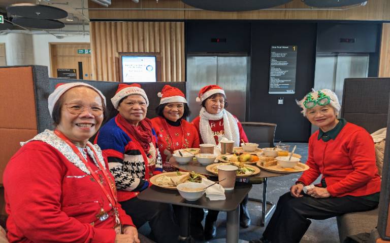 A group of women wearing red jumpers and Santa hats sit around a table with festive party food on inside UCL East's One Pool Street building