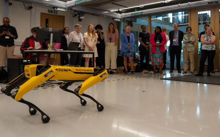 A yellow robotic dog in motion in the foreground, watched by a row of adults in the background in a laboratory setting.