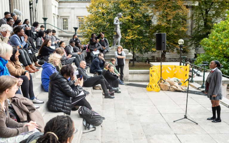 School pupils speaks to an audience sat on the steps of UCL Portico building. 