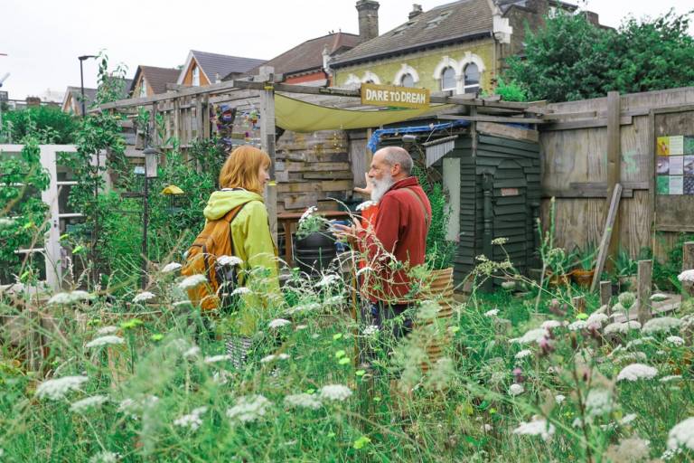 Man and woman talking in front of a shed, in the foreground is grass and flowers