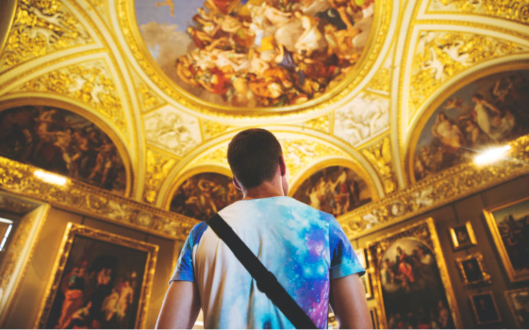 A student looks up at a ceiling painting in an old and grand looking building.