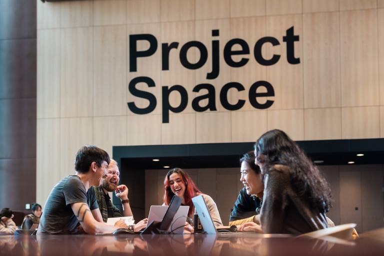 students sitting at a table with laptops talking and laughing below a sign that reads 'project space'
