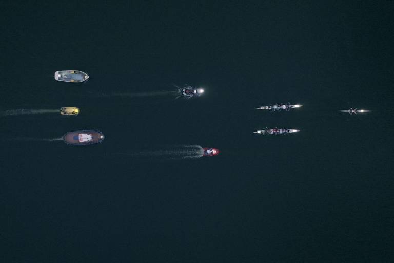 birds eye view of 8 boats travelling from left to right on dark water.