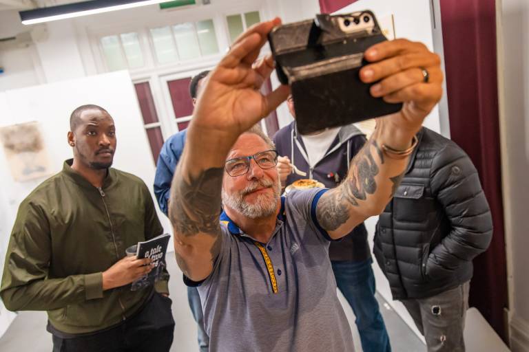 White man with tattoo's is photographing something on their phone in the foreground. Two young Black men look on in the background