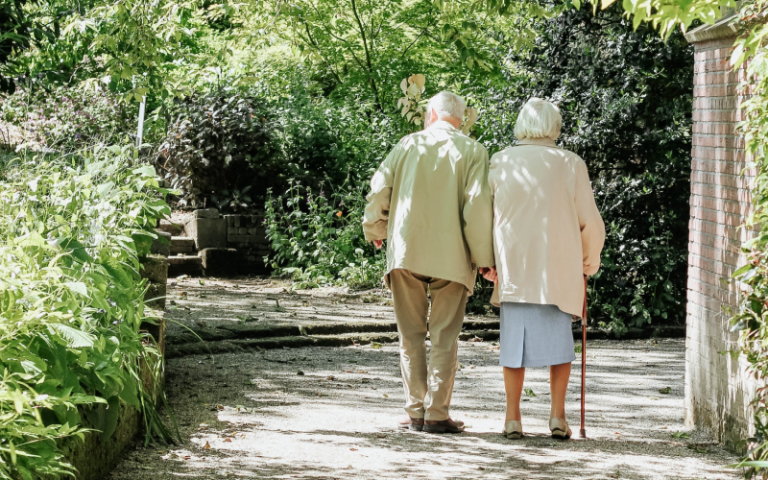 Two elderly people walk together down a wooded path. 