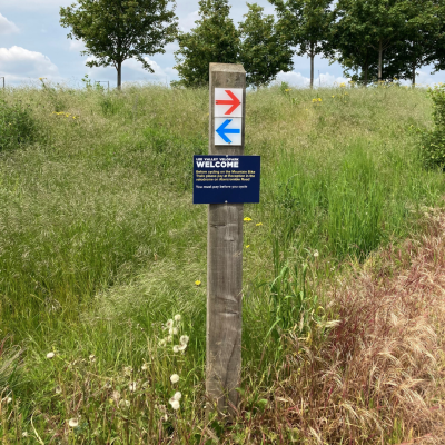 image of wooden sign post sited on grassy lawn with trees on the horizon. On the post are two arrows, one pointing right and one left and text stating Lea Valley Velo Park Welcome.