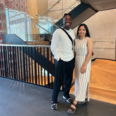 Image of Nate (left) and Chetna (right) standing in front of a spiral staircase in UCL Pool street building. Both are wearing black and white clothes and smiling at the camera
