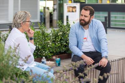 a white man and white woman are sat in conversation on a bench with plants in the background