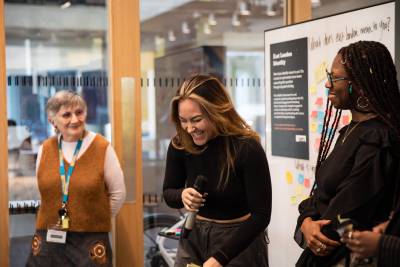 Young black woman is captured laughing whilst holding a microphone, an older white woman looks on. exhibtion boards are in the background