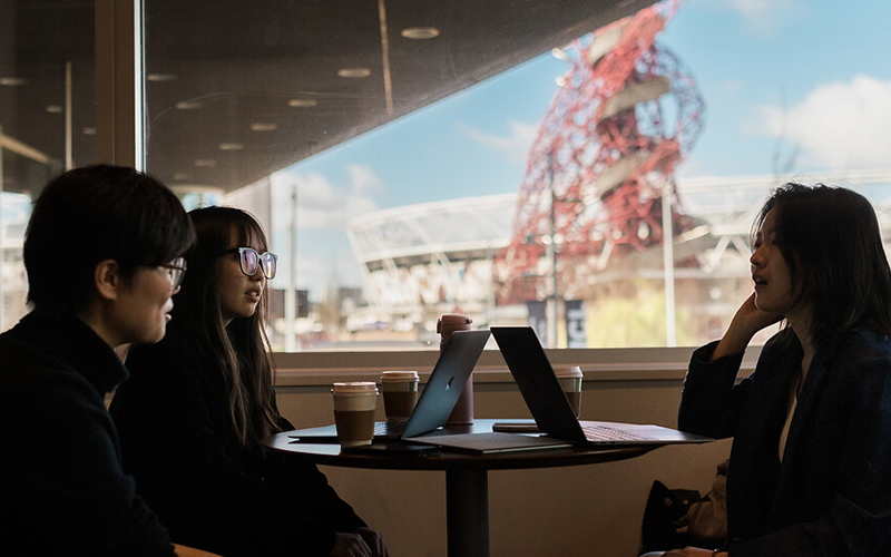 Three students talk with ArcelorMittal Orbit in Queen Elizabeth Olympic Park seen through window