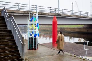 A blurred figure walks past a totem, a  pillar covered in abstract brightly coloured vases