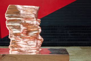 Copper sculpture with undulating surface, in front of red and black wall
