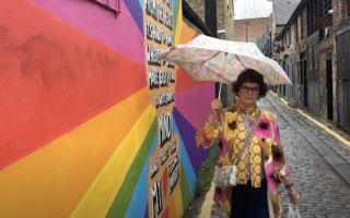 A person stands with a colourful umbrella next to an LGBT rainbow mural.
