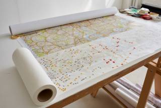 Map of London rolled out on to trestle table with overlaid tracing paper and dots drawn on tracing paper in red, orange and grey ink