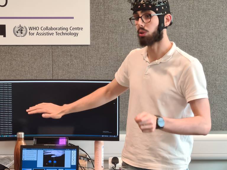 A PhD student wearing a BCI (Brain Computer Interface) cap, is giving a demonstration. His arm outstretched.