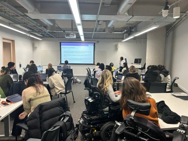 Dr Dilisha Patel teaching the Disability, Design & Innovation MSc. She’s looking at a presentation on a large digital screen in a full classroom.