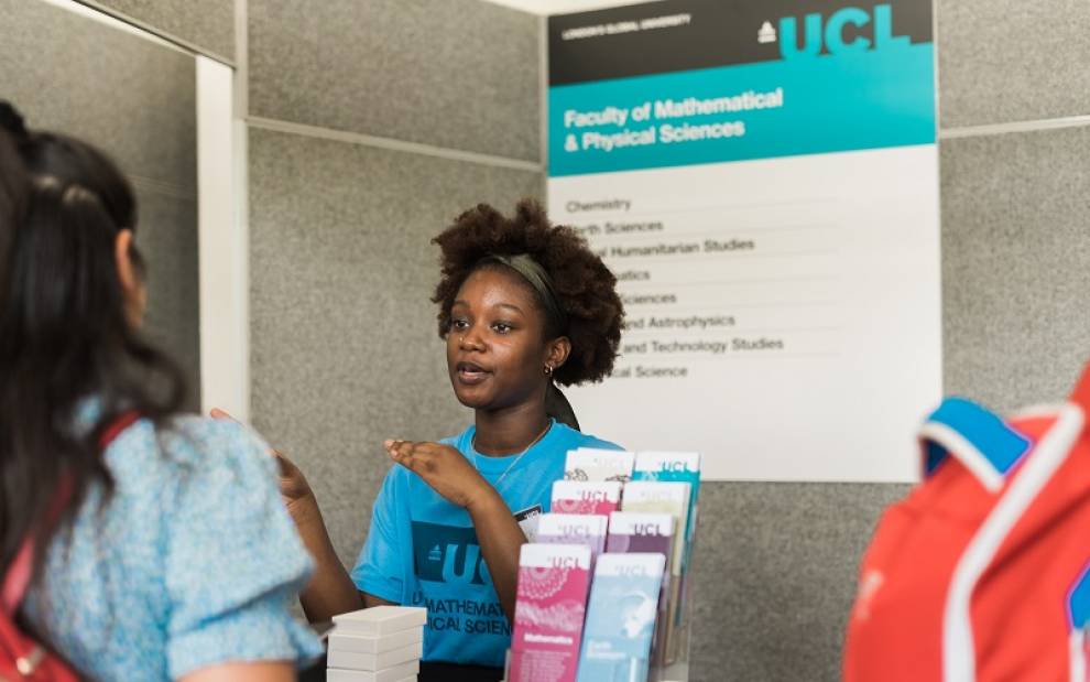 Black femme looking  UCL student wearing a blue T-shirt. She is talking to a visitor at an open day. A sign behind her reads UCL faculty of mathematical sciences