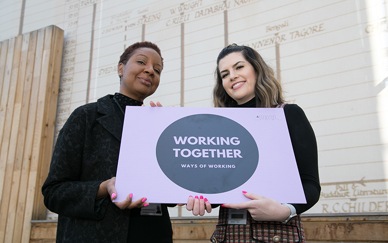 Two members of staff holding up a ways of working - working together sign