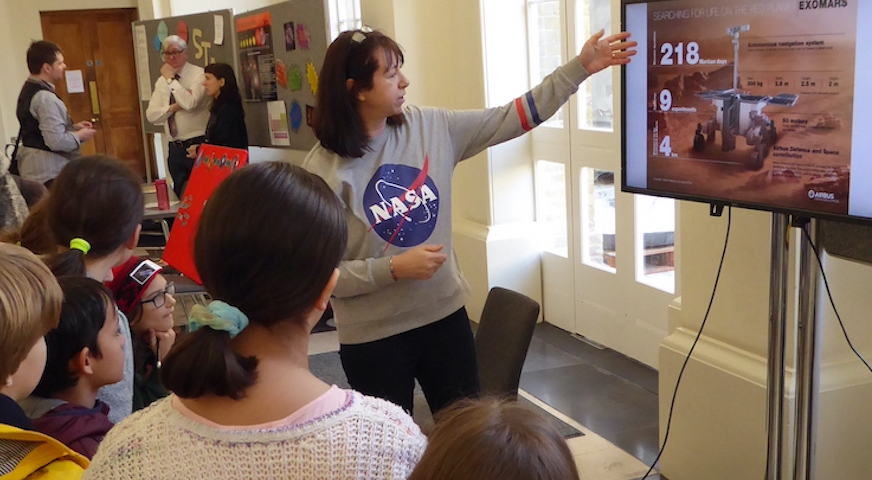 Female with NASA top talking to school pupils in fron of a TV monitor showing a Mars Rover